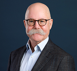 Bald older white male with a gray mustache, and round glasses. Wearing a dark gray blazer and light blue collared shirt