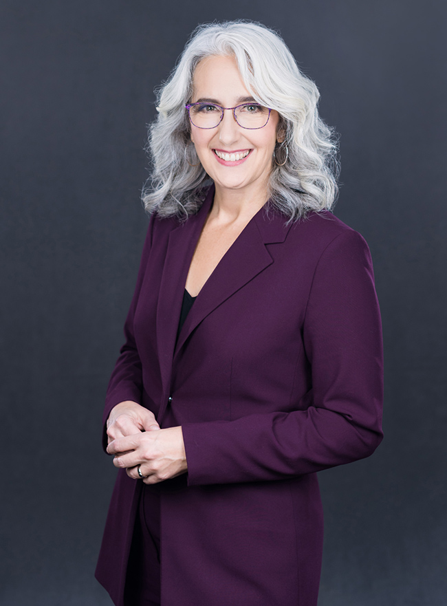 White female with shoulder length gray/white hair, and purple wired framed glasses. Wearing a purple blazer